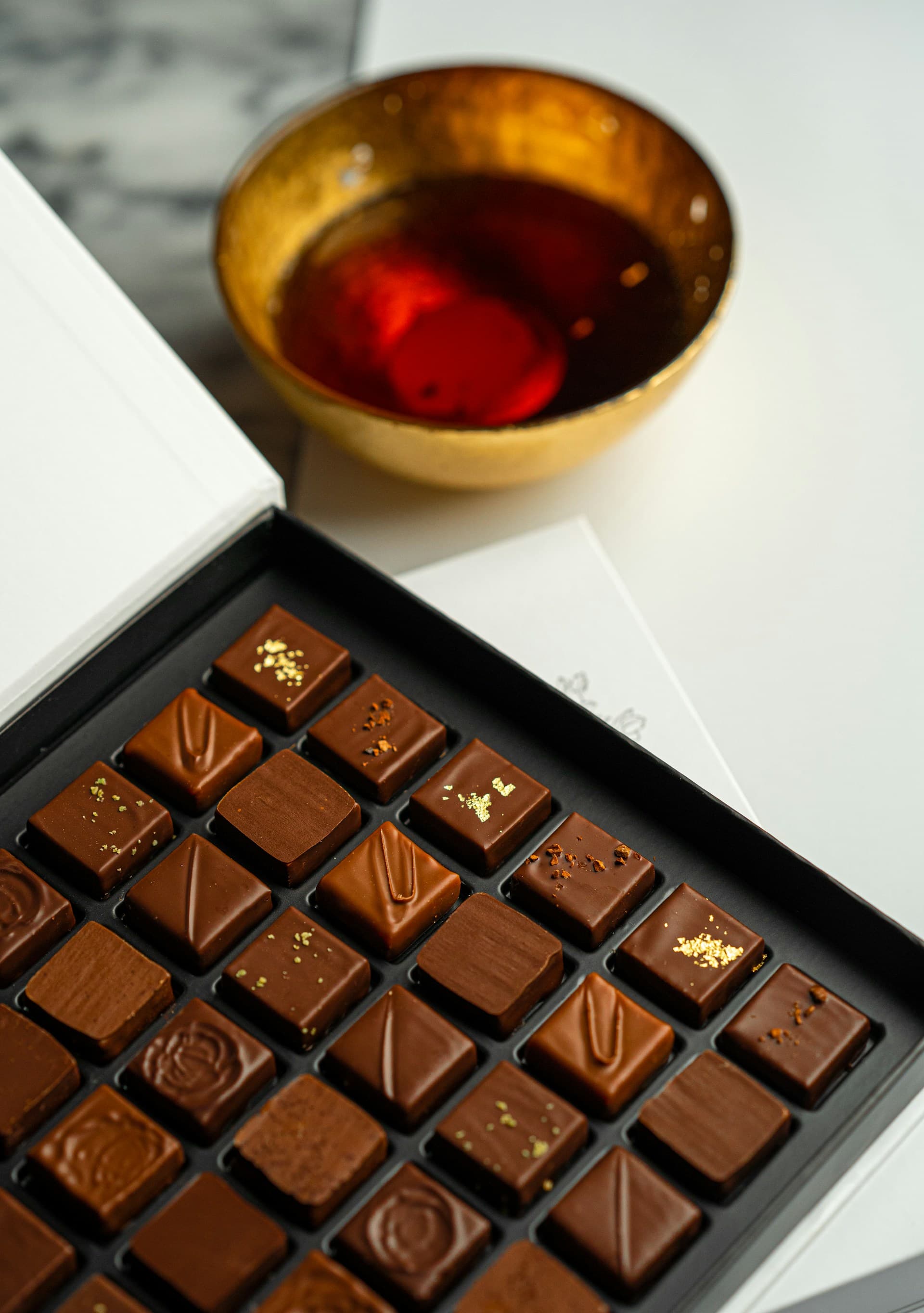 An open box of hand-finished pralines beside a gilded cup of tea